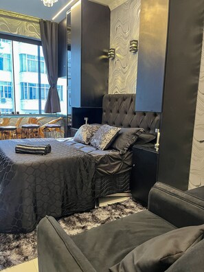 Ein modernes Schlafzimmer mit einem großen Bett, einer Couch, einem Fernseher und Blick auf Gebäude.