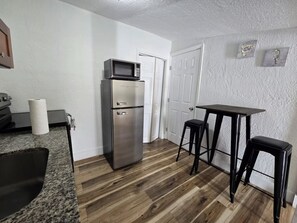 Eine kompakte Küche mit Edelstahlkühlschrank, einer Mikrowelle, Holzboden, einem kleinen Tisch und zwei Barhocker.