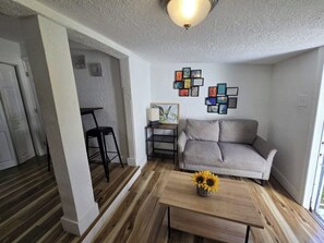 Een woonkamer met een grijze bank, een bijzettafeltje met zonnebloemen, een zwarte fauteuil en een klein tafeltje.