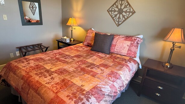 A neatly made bed with a patterned bedspread, two bedside lamps, a small table with a tissue box, and a decorative wall piece. - Oceanfront Suite with Relaxing Tub & Private Bath - Rm 5 (Gabriola)