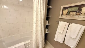 A bathroom with a bathtub, a shower curtain, a towel rack with towels, and a shelf with toiletries. - Oceanfront Suite with Relaxing Tub & Private Bath - Rm 5 (Gabriola)