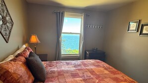 A bedroom with a bed, a nightstand, a lamp, a window with curtains, and a view of the sea. - Oceanfront Suite with Relaxing Tub & Private Bath - Rm 5 (Gabriola)