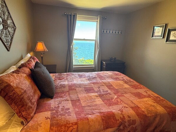 A bedroom with a bed, a nightstand, a lamp, a window with curtains, and a view of the sea.