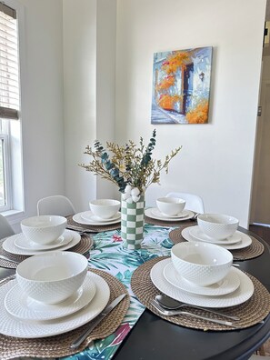 A dining table set with white bowls and plates, a checkered vase with dried flowers, and a painting on the wall. - Across road to Nose Creek Park Cozy city hideaway for family 3BD 2BA/Fenced yard (Calgary)
