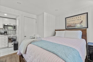 Ein Schlafzimmer mit Bett, einem gerahmten Bild an der Wand, einer Küchenzeile mit Geräten und einem Mülleimer.