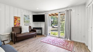 A living room with a brown leather sofa, a flat-screen TV mounted on the wall, a large window with curtains, and a red patterned rug. - Cozy Cape Cod 17A with Fire Pit Near Beaches (Yarmouth)