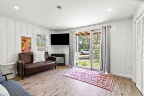 Ein Wohnzimmer mit braunem Ledersofa, einem flachbildfernseher an der Wand, einem großen Fenster mit Vorhängen und einem roten Teppich mit Muster.