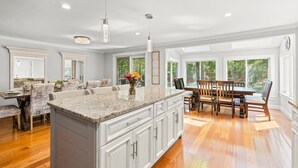 A spacious kitchen with a large island, wooden dining table, and ample natural light from large windows. - Family Getaway | Pickleball Court, Hot Tub and Game Room! (Barnstable)