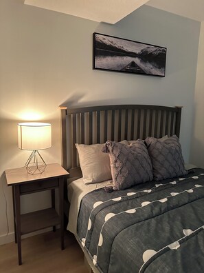 Un dormitorio con cama, mesita de noche con lámpara, cabecero y un cuadro enmarcado en la pared.