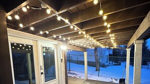 Een overdekt buitengebied met een houten terras, lichtjes en uitzicht op een besneeerd landschap.