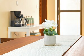 Eine weiße Vase mit weißen Blumen auf einem Holztisch, eine Kaffeemaschine und ein Fenster im Hintergrund.