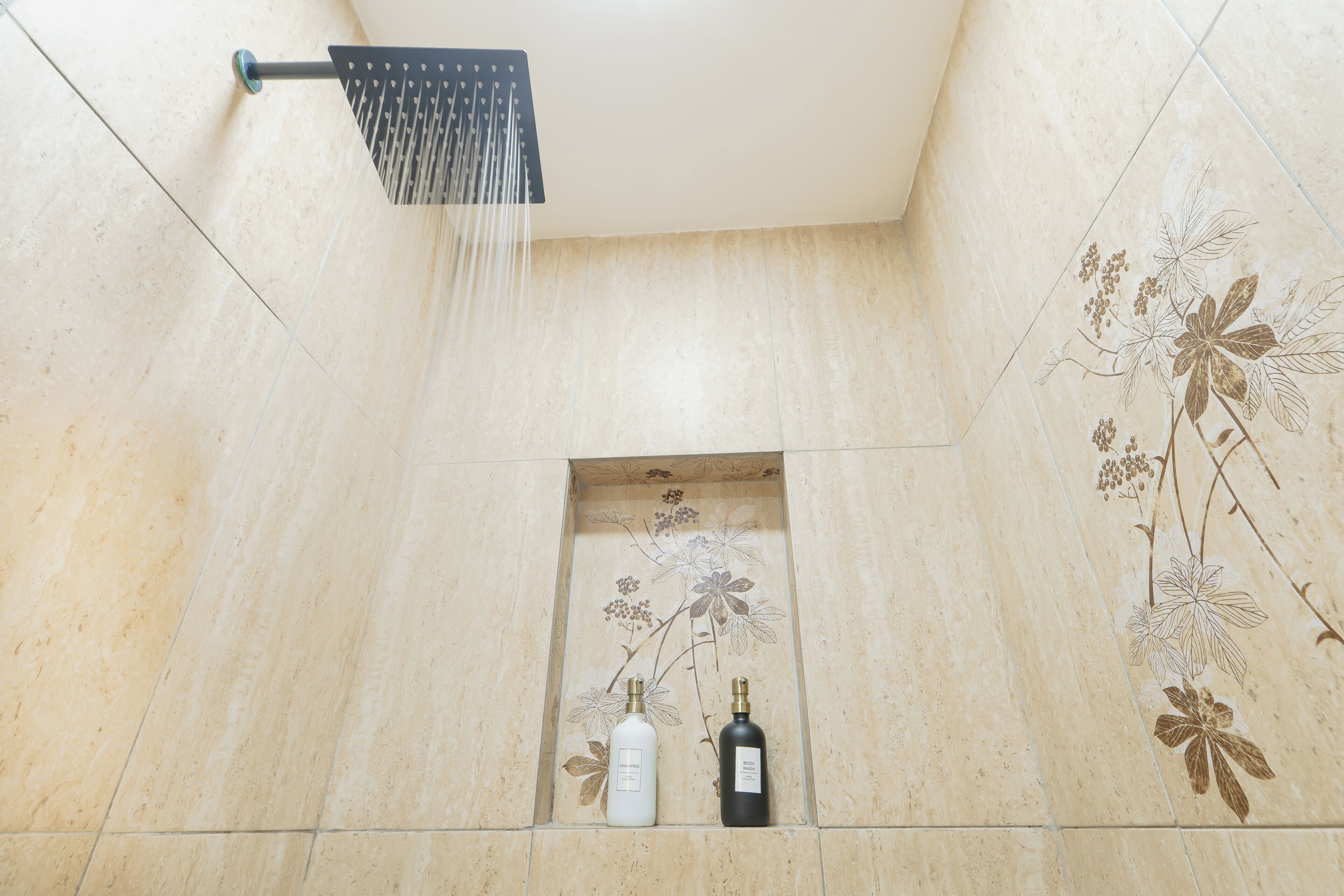 Une douche avec un pommeau de douche en forme de pluie, des carreaux décorés de motifs floraux et deux bouteilles de liquide.