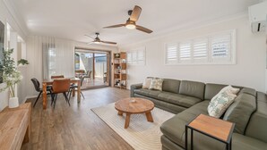 A spacious living area with a sofa, coffee table, and dining table. There are ceiling fans, a bookshelf, and a sliding glass door leading to an outdoor area. - Coastal Comfort with a Private Spa Retreat (Margate)