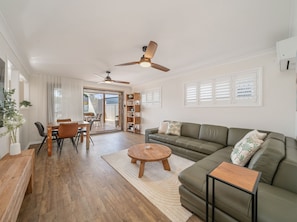A spacious living area with a sofa, coffee table, and dining table. There are ceiling fans, a bookshelf, and a sliding glass door leading to an outdoor area.