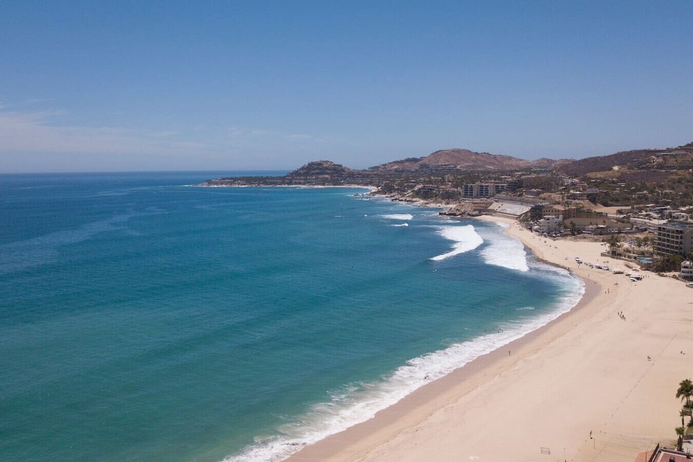 A coastal beach with clear blue waters, a sandy shore, and buildings in the background.