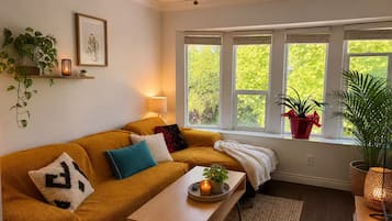 Een gezellige woonkamer met een bank, een bijzettafeltje en een raam met planten.