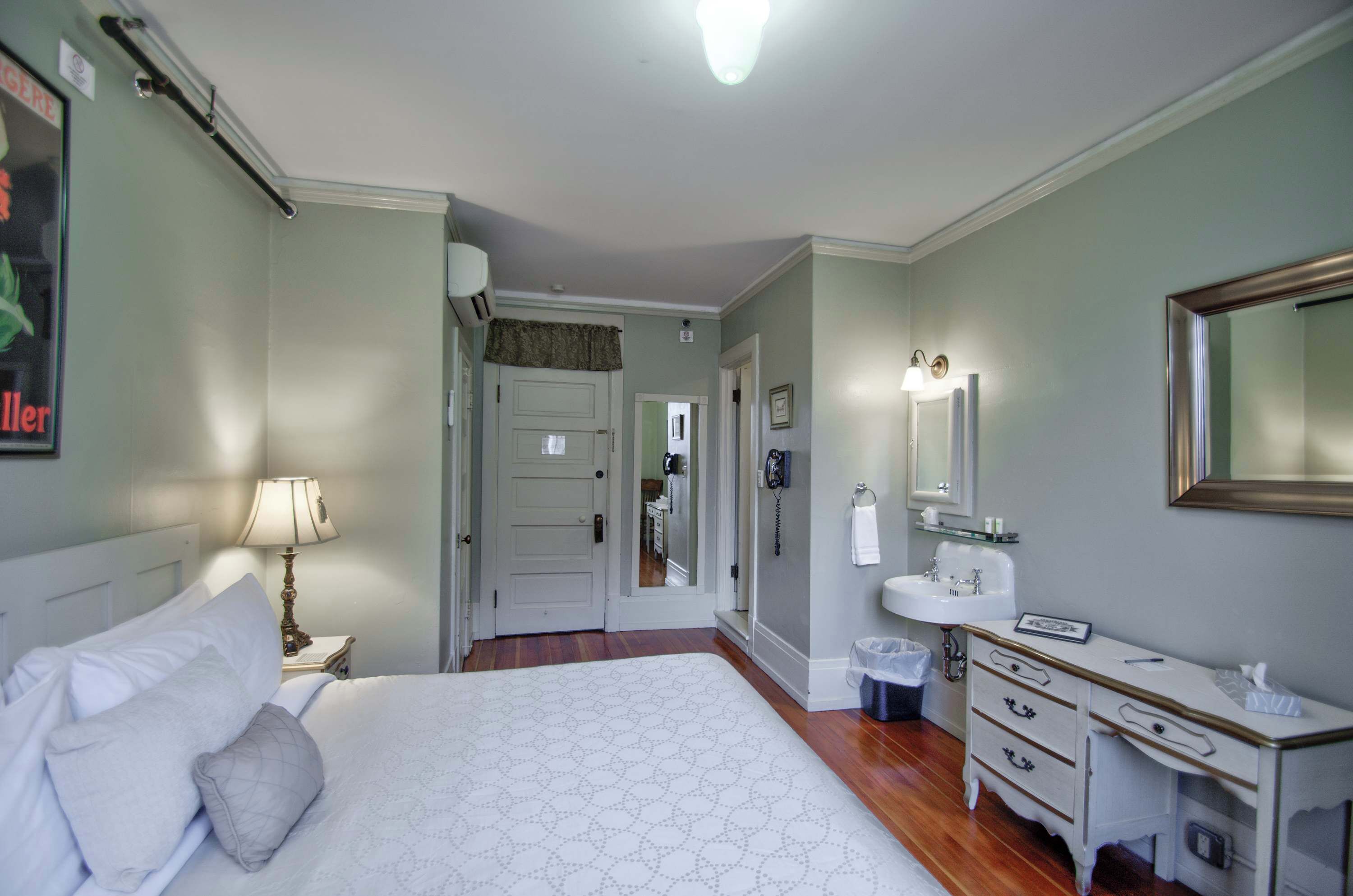 A hotel room with a bed, bedside table, lamp, dresser, mirror, and a bathroom with a sink and mirror.