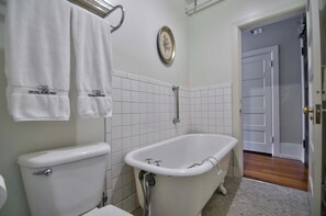 A bathroom with a toilet, a bathtub, white tiled walls, and a door leading to another room. - The Peak at Centralia Square | Historic Suite 201 (Centralia)
