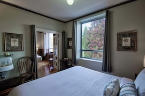 A bedroom with a large bed, a window with a view of trees, a chair, a small table, and a sink.