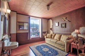 Una habitación con sofá, una mesita con lámpara, espejo, ventana con vistas y alfombra estampada.