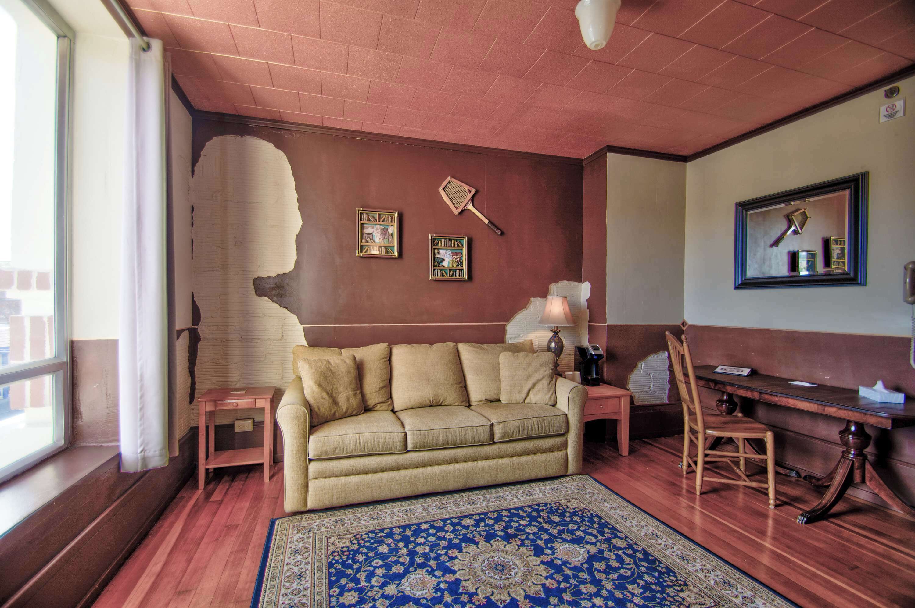 A room with a wooden floor, a beige sofa, a small table, a lamp, a chair, and a blue patterned rug.