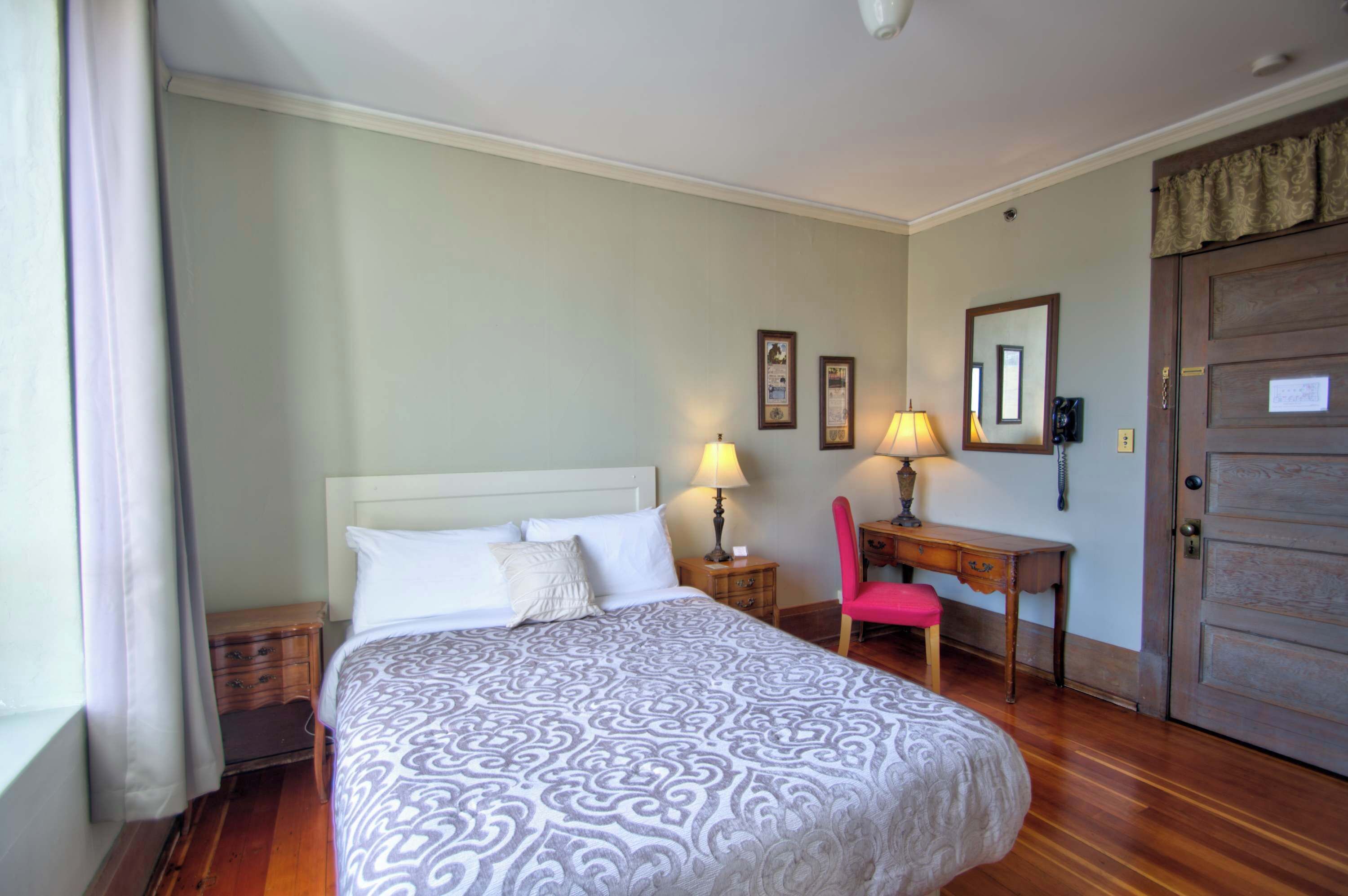 A hotel room with a bed, two bedside tables, a desk with a chair, a door, and a window with curtains.