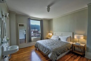 Un dormitorio con cama, lámparas de noche, ventana con cortinas, puerta y baño con lavamanos.