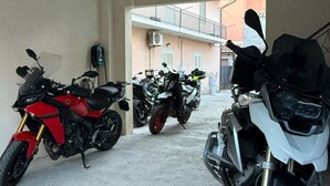 A covered motorcycle parking area with three motorcycles, a concrete floor, and a building in the background. - Old Town Room 1 (Tortolì)