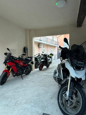 Area coperta per il parcheggio di motociclette, con tre moto, pavimento in cemento e un edificio sullo sfondo.