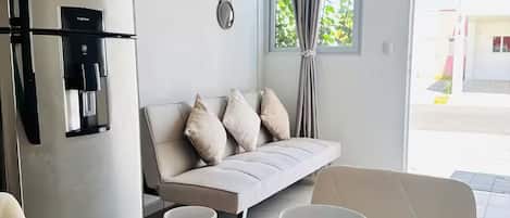 A modern living room with a sofa, dining table set with plates, cups, and cutlery, a refrigerator, and a window with curtains.