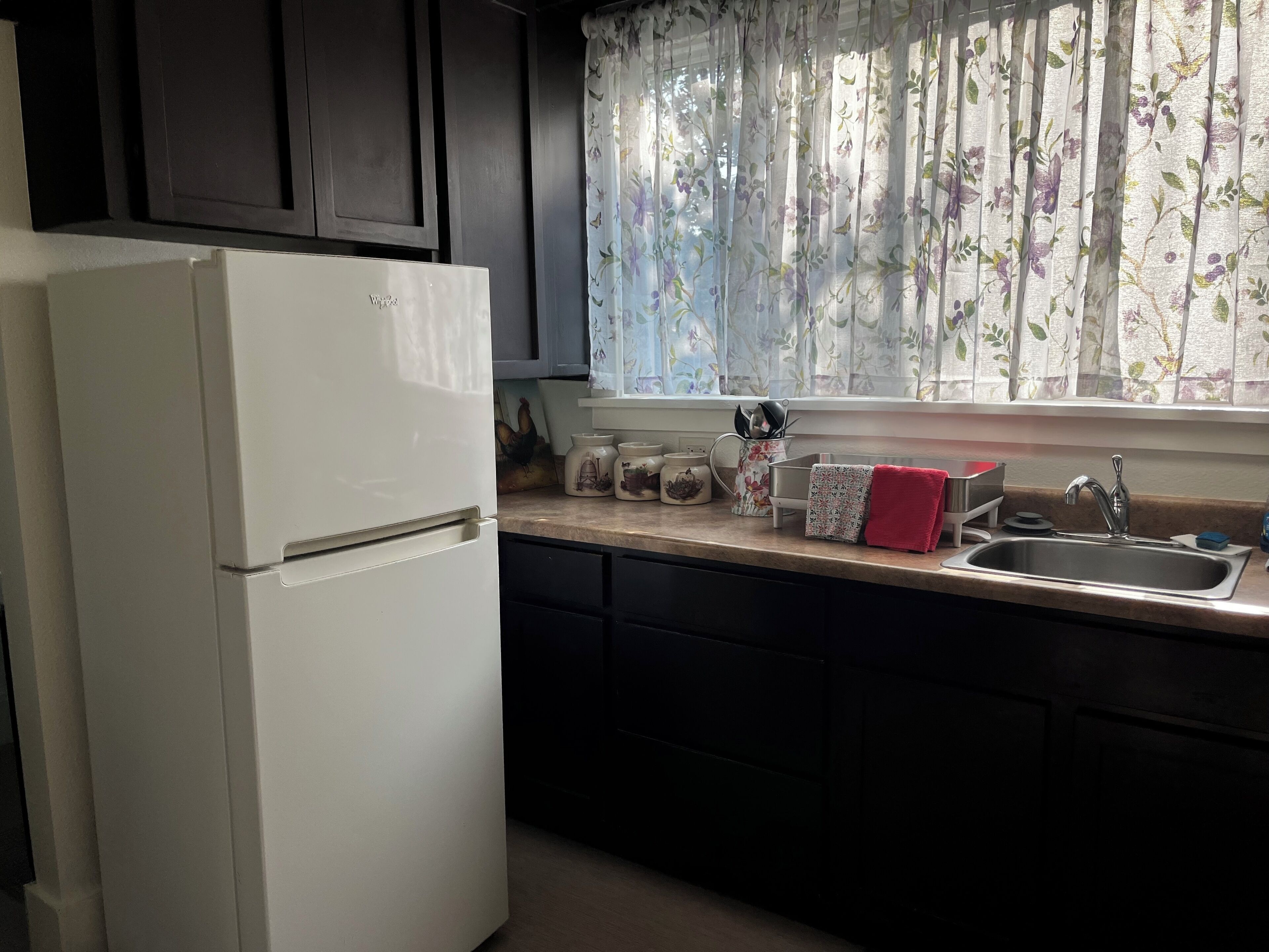 A kitchen with a white refrigerator, dark cabinets, a window with floral curtains, and a stainless steel sink.