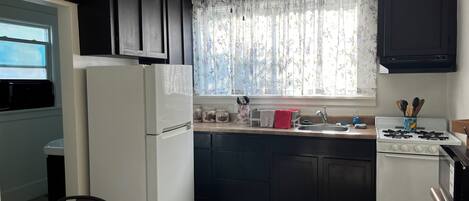 A compact kitchen with a refrigerator, stove, sink, and window with floral curtains.