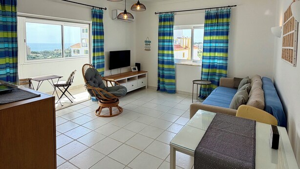 A modern living room with a sofa, dining table, and chairs, a TV, and a view of the sea. - Azul,: A cozy apartment just a few steps from Praia da Rocha (Portimão)