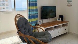 A room with a wicker chair, a TV on a wooden stand, a curtain, and an air conditioner. - Azul,: A cozy apartment just a few steps from Praia da Rocha (Portimão)