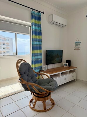 A room with a wicker chair, a TV on a wooden stand, a curtain, and an air conditioner. - Azul,: A cozy apartment just a few steps from Praia da Rocha (Portimão)