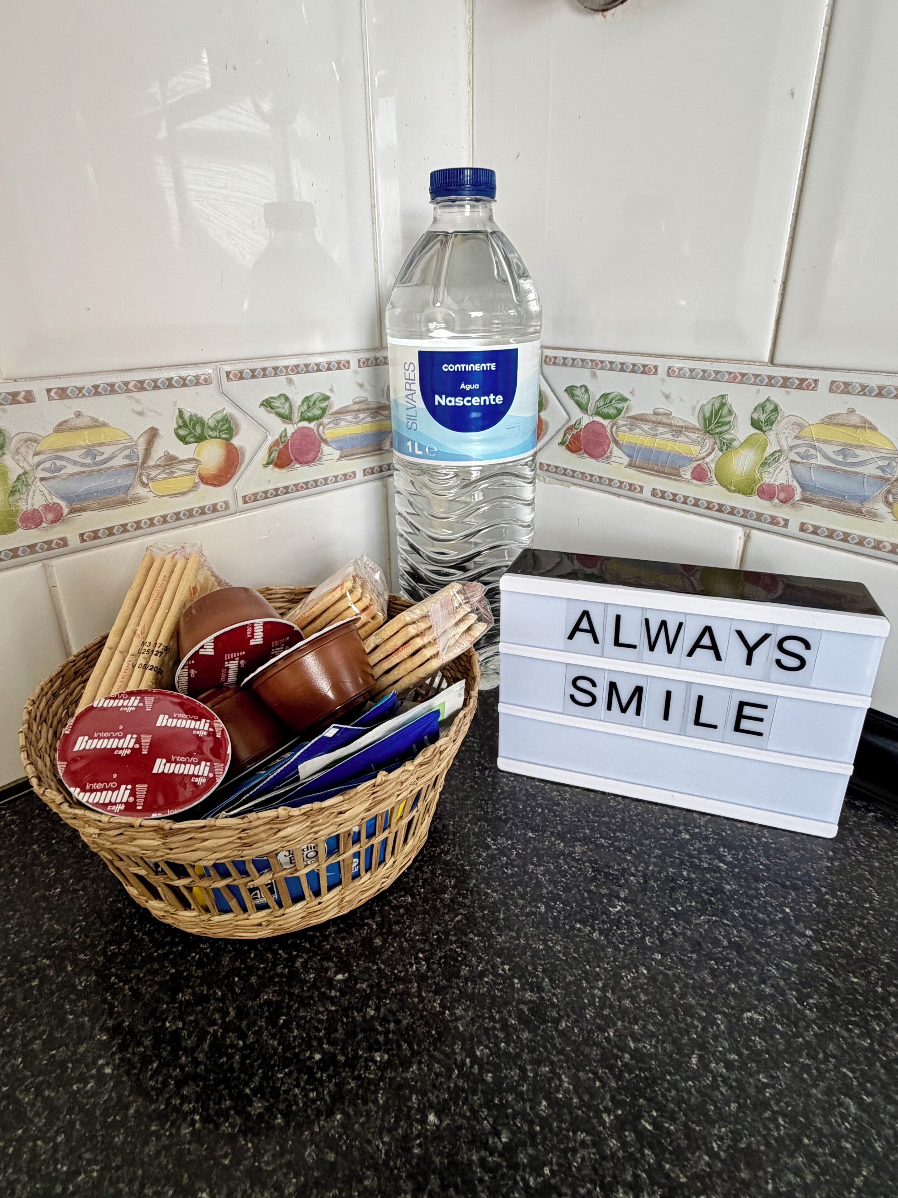 A kitchen counter with a water bottle, a basket of snacks, and a sign that says 'ALWAYS SMILE'.