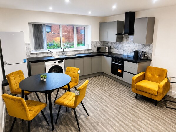 A modern kitchen with a washing machine, a dining table with chairs, and a view of a brick building. - Modern 2-Bed Flat Near Donington & Airport (Long Eaton)