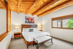 Ein Schlafzimmer mit Holzbalkendecke, einem großen Bett, Nachttischlampen, einem Gemälde von einer Kuh und einem Fenster mit Blick.