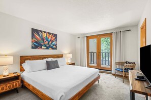 Ein Hotelzimmer mit Bett, zwei Nachttischen, einem Fernseher, einem Sessel und einem Balkon mit Vorhängen.