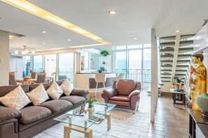 A modern living room with a sofa, armchair, and glass coffee table. A staircase with a statue is visible in the background. - The Cebu Skyline Penthouse Romantic Getaway with Pool, King size Bed, 2 BR (Cebu City)