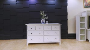 Une commode blanche avec de nombreux tiroirs, un vase rempli de fleurs et un mur noir orné de motifs géométriques.