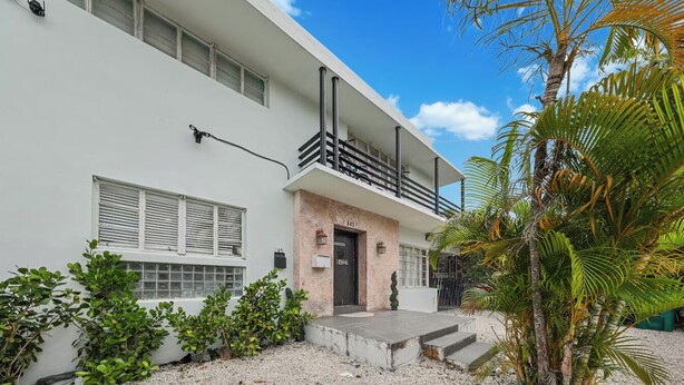Un edificio moderno con un balcone, una porta con un numero civico e un vialetto ghiaioso.