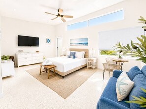 Un dormitorio moderno con cama, mesitas de noche, televisor, ventilador de techo, ventana y vistas al exterior.