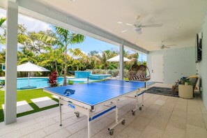 Ein Tischtennis-Tisch in einem überdachten Außenbereich mit Pool und Palmen im Hintergrund.