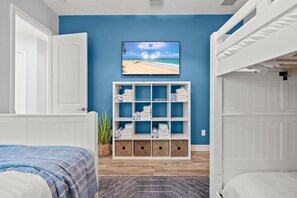 Ein Schlafzimmer mit blauer Wand, einem Fernseher, der über einer weißen Schrankwand angebracht ist, und einem Bett mit einer blau-weißen Streifenauflage.