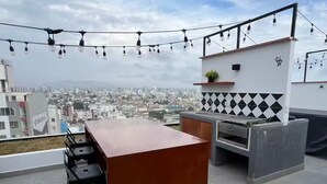 Un bar situé sur le toit, offrant une vue sur la ville, avec un comptoir, des tabourets, un barbecue et des guirlandes lumineuses.