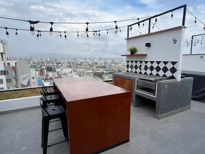 Un bar en la azotea con vista a la ciudad, una barra, taburetes, una parrilla y luces colgantes.