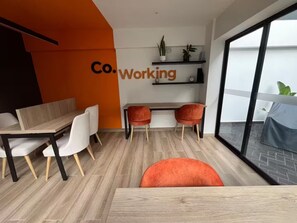 Un espacio de oficina moderno con una mesa de madera, sillas blancas y una silla roja de acento. Se observa una estantería con plantas.