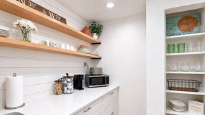 Een keuken met witte kastjes, een roestvrijstalen spoelbak, houten planken met keukengerei, een magnetron en een afvalbak.