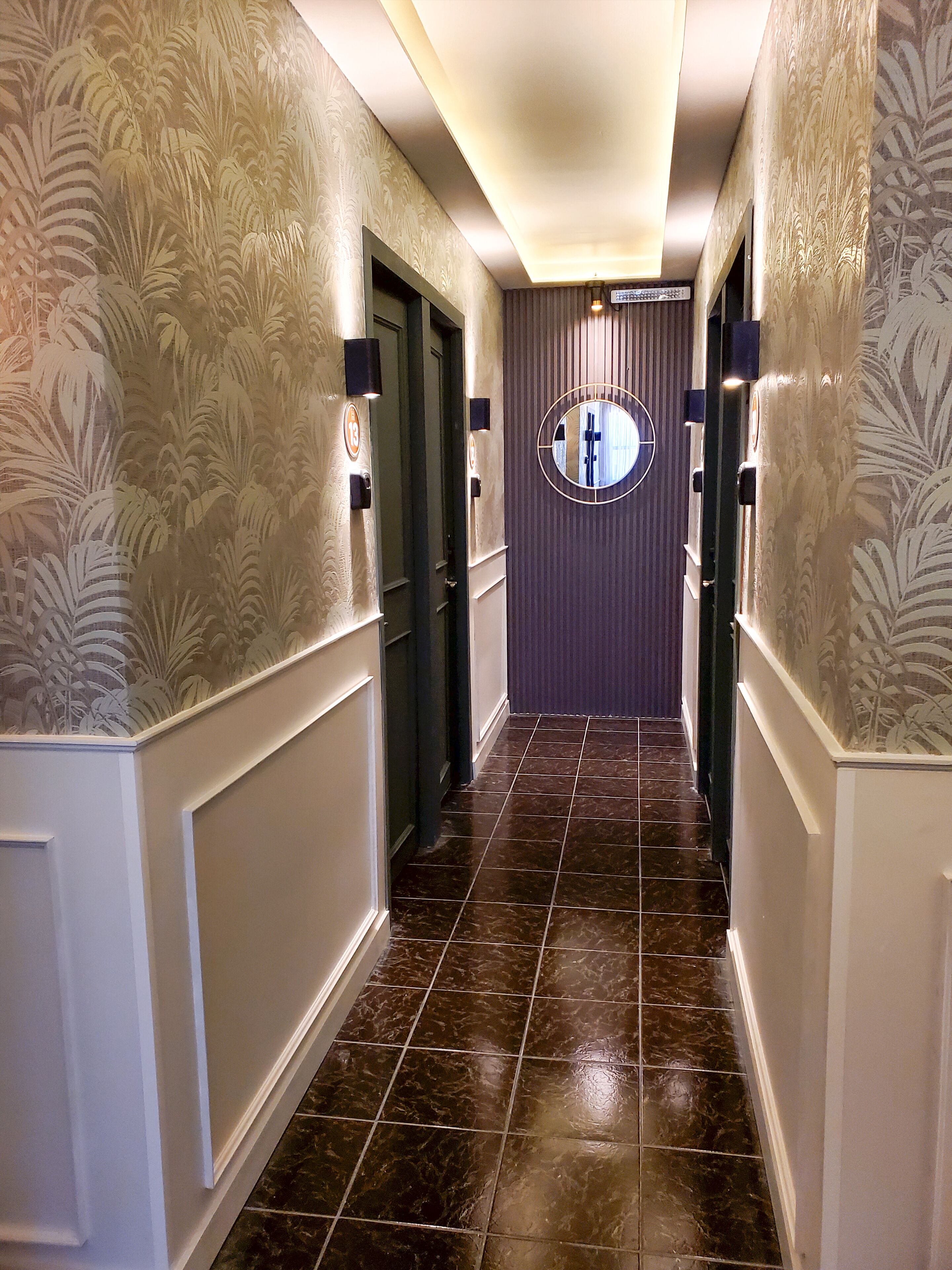 A hallway with patterned wallpaper, dark tiled flooring, and a circular mirror on the wall.